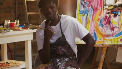 Portrait-of-african-american-male-artist-wearing-apron-holding-a-paint-brush-smiling-at-art-studio