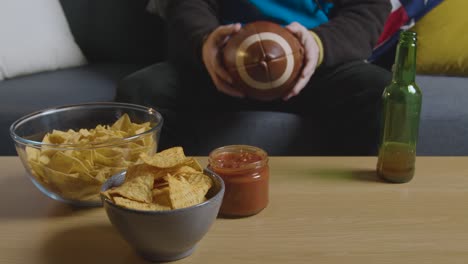 person celebrating at home watching american football game on tv with drinks snacks and ball