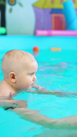 lovely smiling baby moved in water by his coach. female trainer shows how to breath in swimming pool. vertical video.