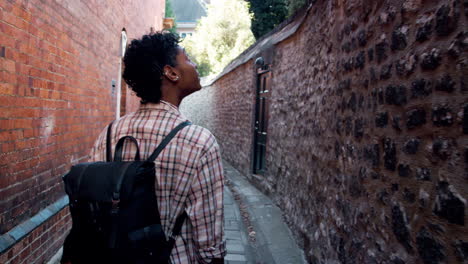 Millennial-black-woman-wearing-a-plaid-shirt-carrying-a-backpack-walking-in-a-narrow-alleyway-in-a-historical-district-and-looking-around,-back-view,-follow-shot