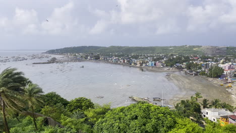 a breathtaking aerial drone view of uttan beach in mumbai, maharashtra, featuring a cloudy sky, birds soaring, lush green trees lining the coast and the cityscape into the horizon
