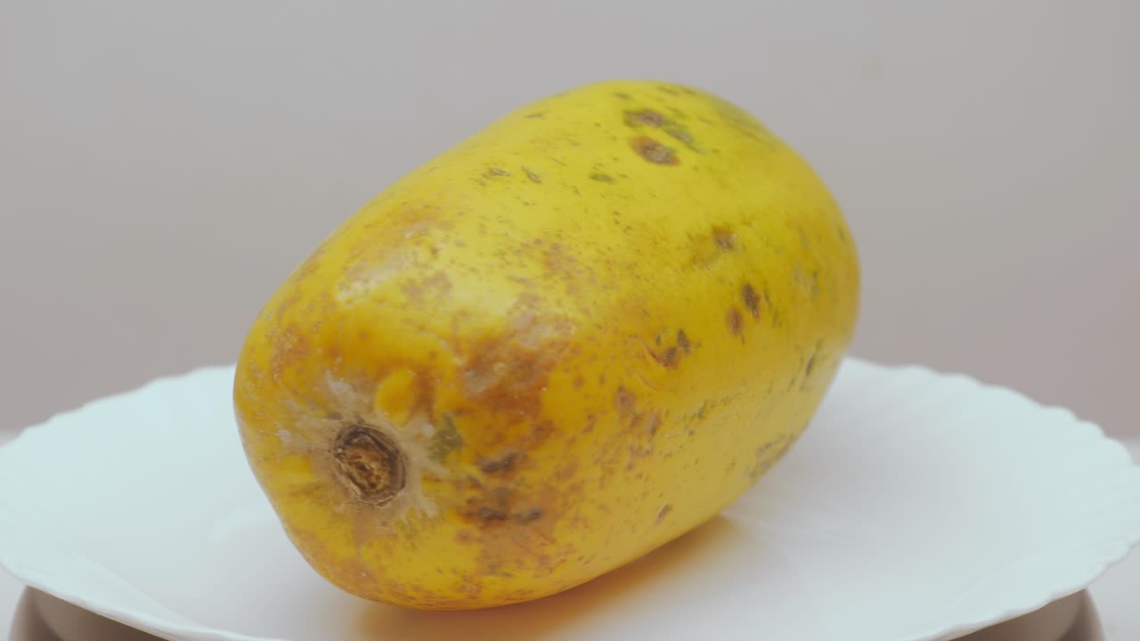 A single ripe papaya slowly rotates on a white display plate, showing its warm yellow skin, natural blemishes, and smooth elongated shape under soft studio lighting