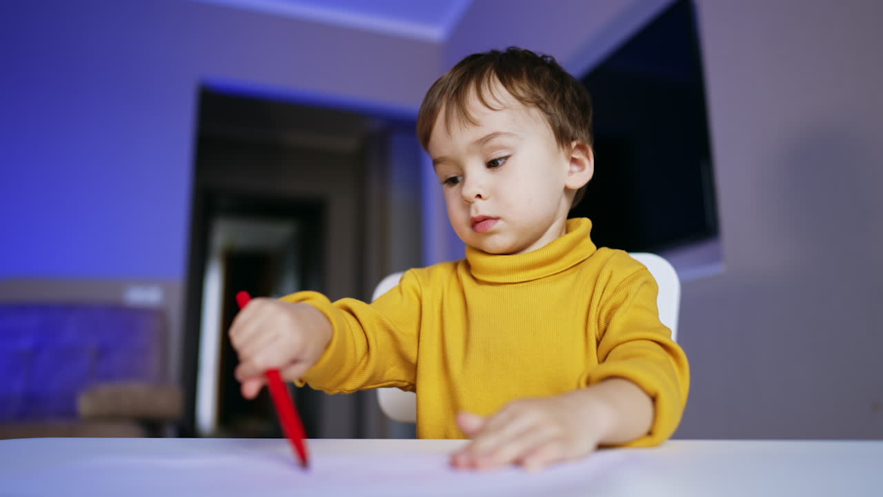 Adorable dark-haired baby boy drawing peacefully at home. Lovely toddler in yellow sweater uses red pen. Low angle view.