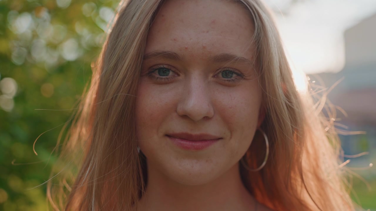 Young woman with long blonde hair smiles and speaks directly to camera in soft natural light her expression changing from calm conversation to open mouthed laughter. background features