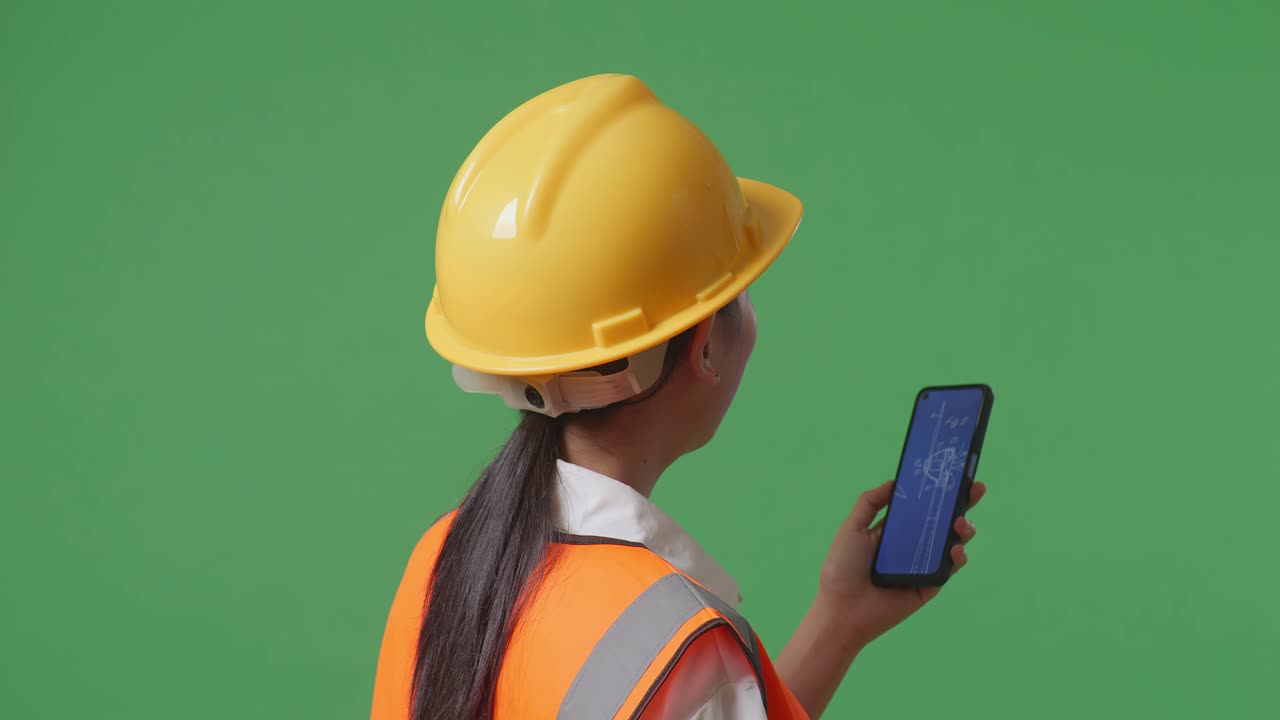 안전 헬을 입은 아시아 여성 엔지니어가 녹색 화면 배경 스튜디오에 서 있는 동안 스마트폰에서 풍력 터빈 블루프린트를 보고 있는 가까운 뒷면