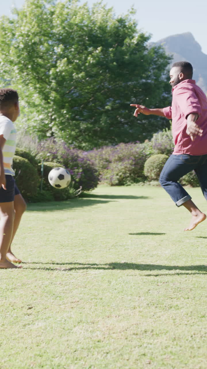 Vertical video of african american father and son playing football in garden, in slow motion
