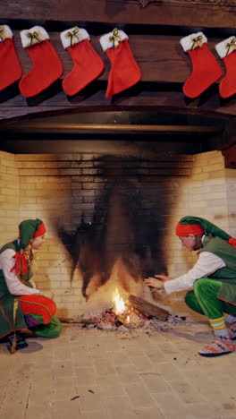 christmas background with two elves sitting near the burning fireplace at living room. red socks hanging over the fireplace and fairy elves warming up their hands close to the fire indoors. vertical video