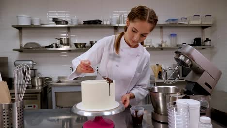 un chef de pastelería vertiendo chocolate líquido de una cuchara en un pastel de esponja de crema blanca