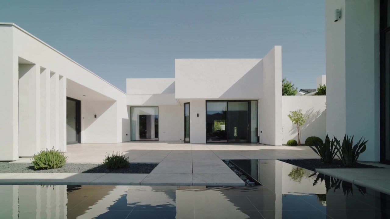 La cámara se aleja, mostrando la fachada de una casa blanca en un patio, con una piscina reflectante para realzar su forma.