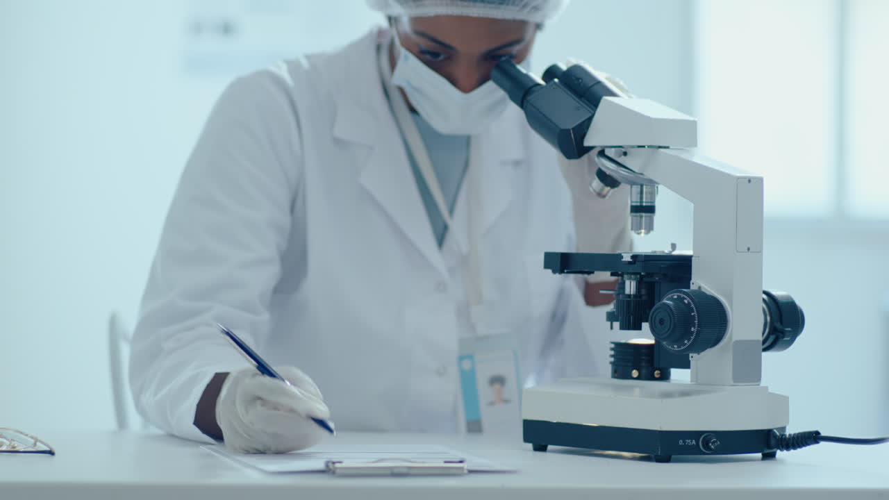 African-American Female Scientist Using Microscope and Making Notes in Lab