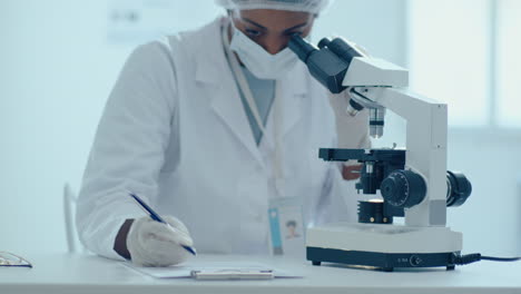 científica afroamericana usando el microscopio y tomando notas en el laboratorio