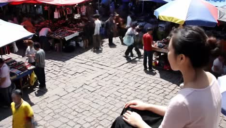 a young woman observes a lively market in an ancient town where vendors display colorful crafts and visitors browse through various goods under bright umbrellas on a sunny day.