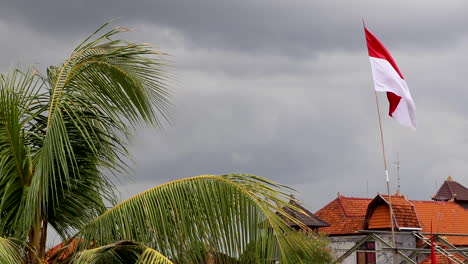 Merah-Putih,-Bandera-Indonesia-Ondeando-Contra-Nubes-Tormentosas-Con-Palmeras-Y-Tejados-Tradicionales