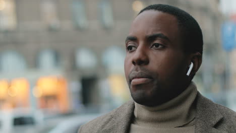 Close-up-view-of-african-american-businessman-with-a-beard-wearing-wireless-headphones-while-talking-on-the-phone-in-the-street-in-autumn