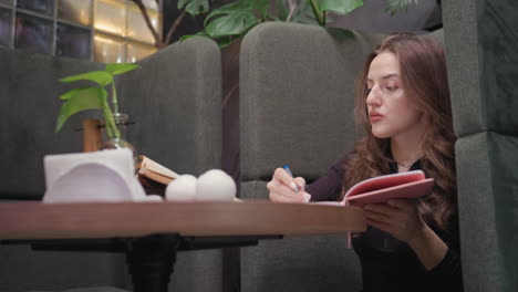 lady in black outfit calmly opens pink notebook and begins writing while seated in green booth. scene includes decorative plant and open brown book on table, with peaceful indoor greenery around her