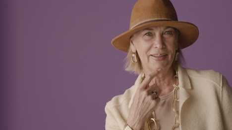 Blonde-Mature-Woman-With-Blue-Eyes-Dressed-In-Jacket,-Hat-And-Accessories-Posing-With-Finger-On-Chin-And-Smiling-At-Camera-On-Purple-Background