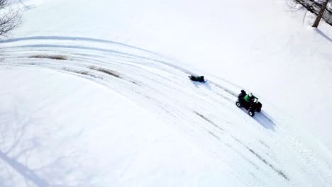 A-drone,-aerial,-birds-eye-top-down-view-of-an-atv-4-wheeler-with-a-child-riding-on-the-back-and-pulling-another-child-riding-on-their-belly-on-a-winter-sled-on-the-snow-covered-ground-in-the-country