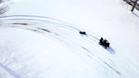 a drone, aerial, birds eye top down view of an atv 4 wheeler with a child riding on the back and pulling another child riding on their belly on a winter sled on the snow covered ground in the country