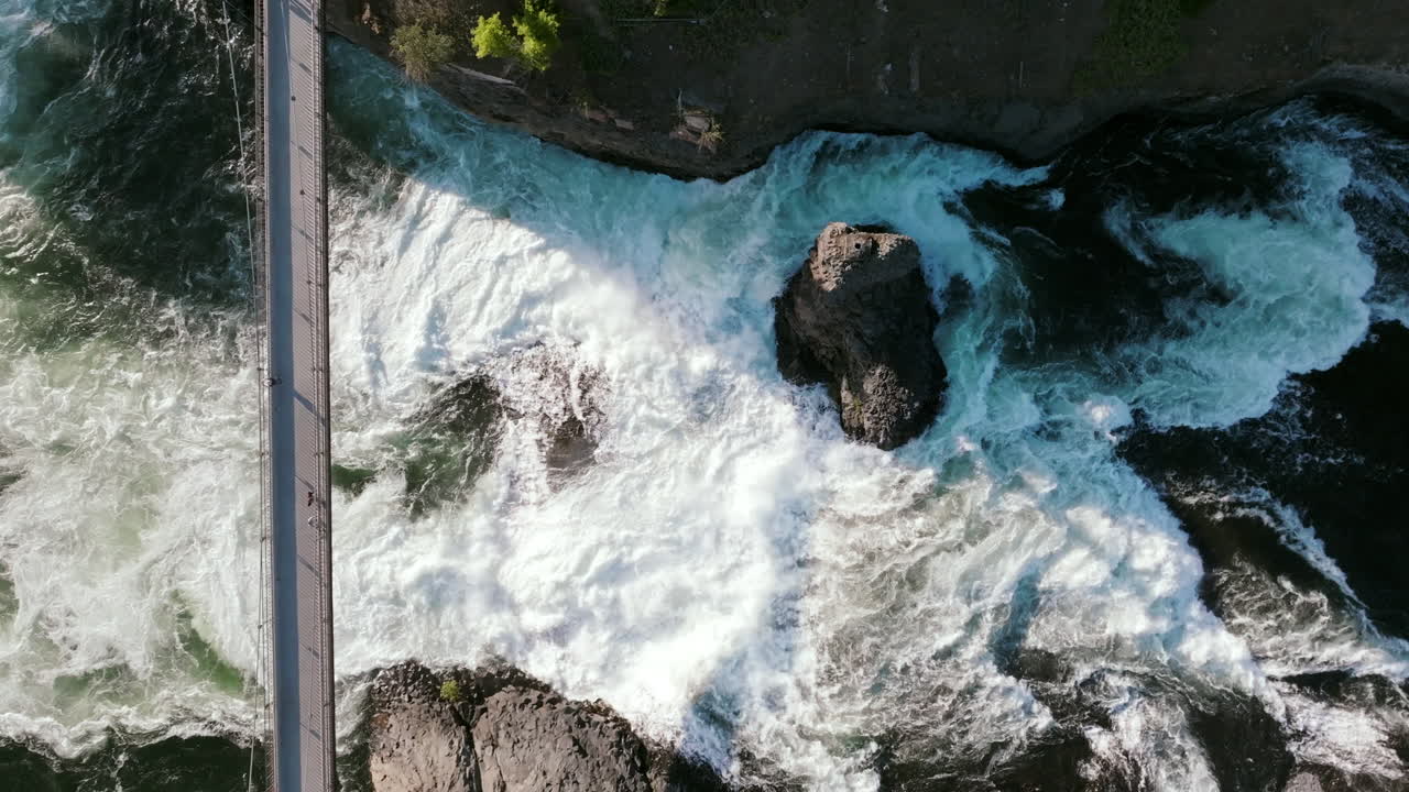 Whitewater crashes around rocks below a pedestrian bridge. The river flows fast and loud through the middle of the city