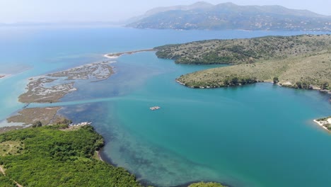 Drone-view-in-Albania-flying-over-wide-river-and-green-landscape-next-to-the-sea-with-mountains-on-the-back