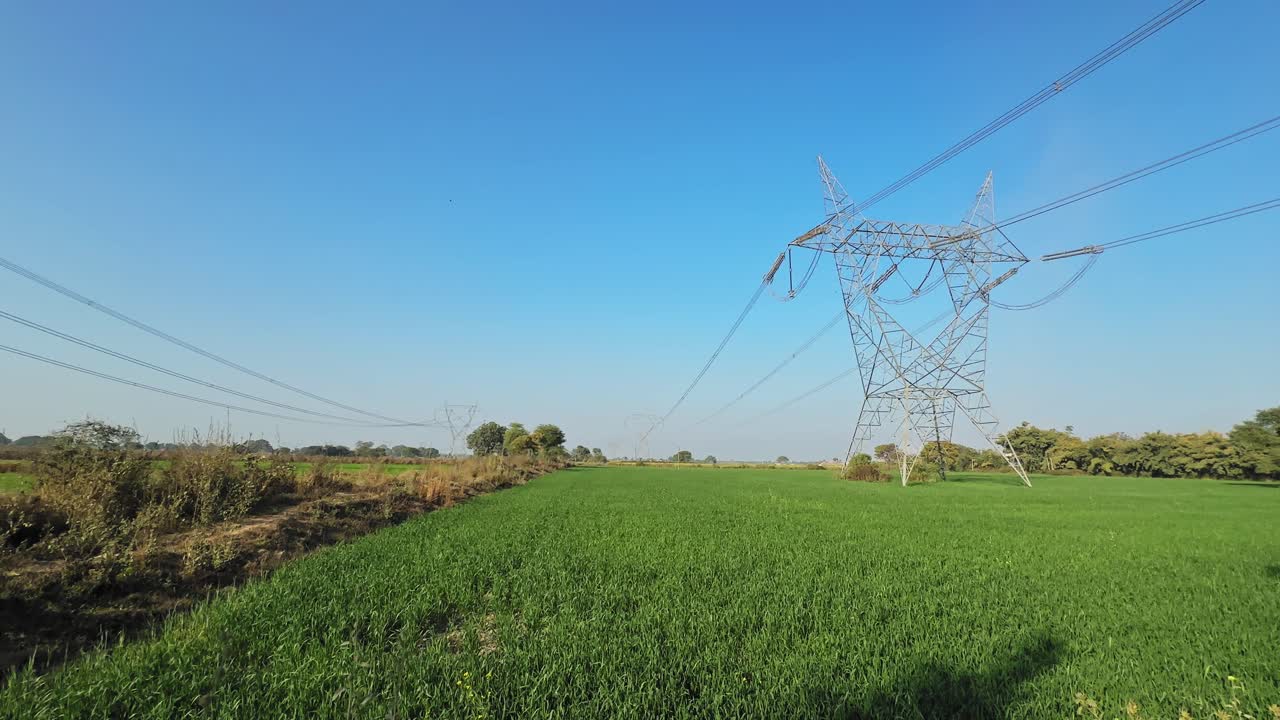 Panning shot of high voltage transmission towers and power lines stretching across green agricultural fields under a clear blue sky in an open rural landscape