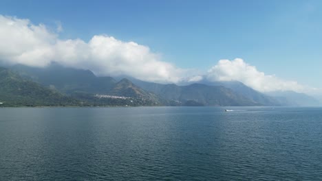 drone view in guatemala flying over a blue lake surrounded by green mountains approaching a moving boat on a sunny day in atitlan