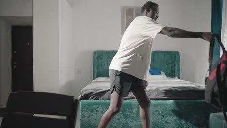 man in white t-shirt holding backpack, walking through minimalist bedroom, with green headboard, bed with neatly folded towels, and natural light coming from the window