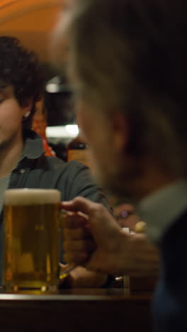 vertical shot of young bartender pours craft beer into glass and gives it to mature man. male friends clink glasses and drink beer sitting at the bar counter in stylish pub at night. concept of lifestyle and leisure.