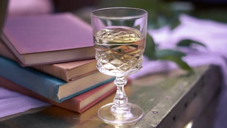 a rearward bokeh shot of a diamond-styled glass of white wine beside a stack of colorful books and leaves on top of a white cloth on a wooden table