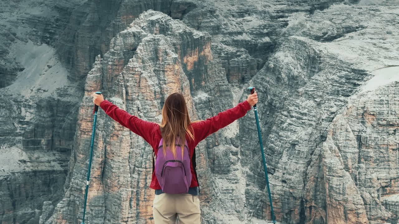 Female hiker with backpack and trekking poles opening arms facing the rocky peaks of the Dolomites, Italy