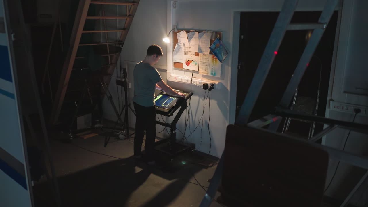 Young man operates touchscreen control panel in low-light studio near wall with messy notes, diagrams, and plugged-in cords while surrounded by technical equipment