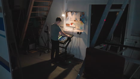 young man operates touchscreen control panel in low-light studio near wall with messy notes, diagrams, and plugged-in cords while surrounded by technical equipment