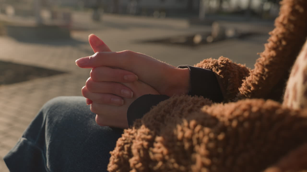 una toma de primer plano de una persona sentada con su cara no visible, con pantalones vaqueros azules y un abrigo marrón. sus manos están apretadas fuertemente, sugiriendo un momento de pensamiento profundo, meditación
