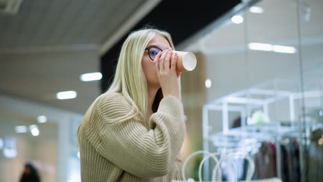 young woman with glasses sipping beverage while sitting in mall with shopping bags, she looks contemplative, with a blurred background featuring a clothing store and other shoppers walking