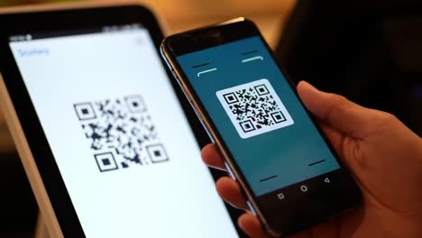 a person uses a smartphone to scan a qr code displayed on a tablet, facilitating a seamless payment process at a popular cafe. the atmosphere is busy, filled with customers enjoying their beverages.