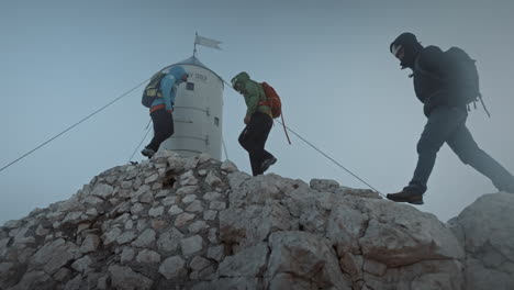 Hikers-with-backpack-and-helmets-arriving-at-the-peak-of-mountain-Triglav-in-front-of-Aljaž-Tower,-sky-is-clear