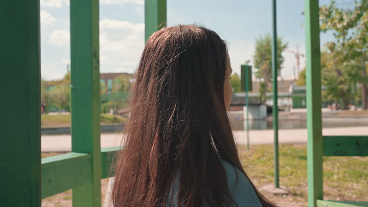 따뜻한 햇살 아래 평화로운 공원을 배경으로 파란색 블라우스를 입고 녹색 울타리 옆에 조용히 서 있는 여성의 뒷모습이에요. 게이트 근처에 있는 한 백인 여성이 머리카락을 만지고 있어요.