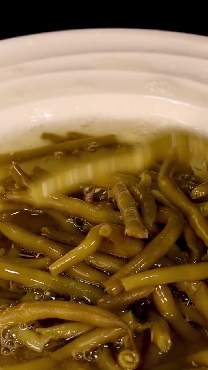 A haricot bean is dropped into a white bowl filled with cooked green beans and liquid, causing a splash. Close-up, warm lighting, static camera