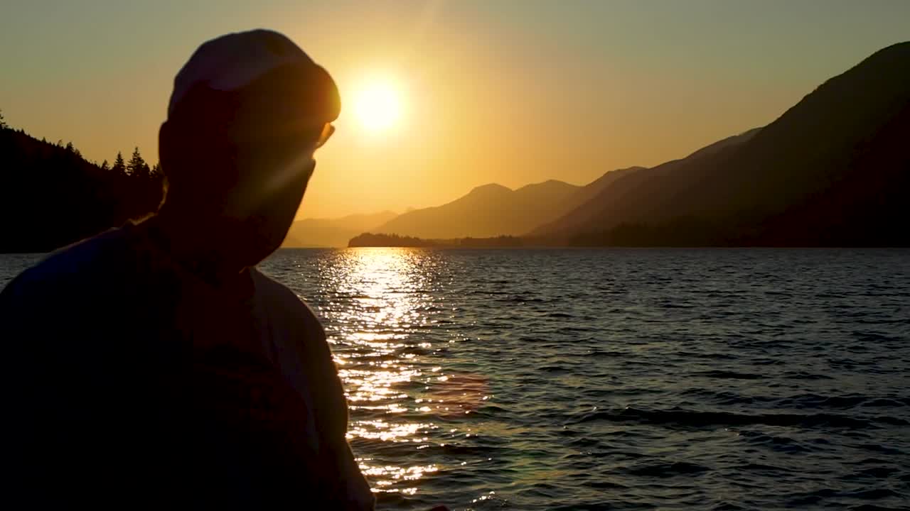 Tramonto su un lago con montagne sullo sfondo, con un ragazzo che controlla il telefono in controluce.