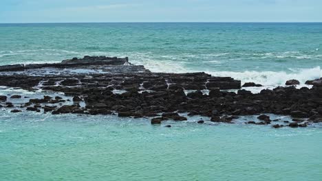 aerial pan of low rocky outcropping as ocean water splashes from waves on basalt plains