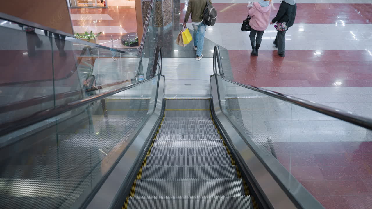 Back view of shopper in green jacket carrying shopping bags descending escalator in spacious mall with glass railing reflections bright tiled floor and clean architectural symmetry