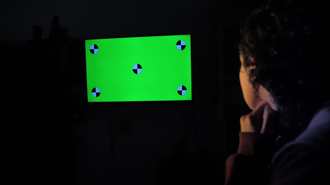 Persona che guarda una TV con schermo verde e marcatori di tracciamento in una stanza buia.