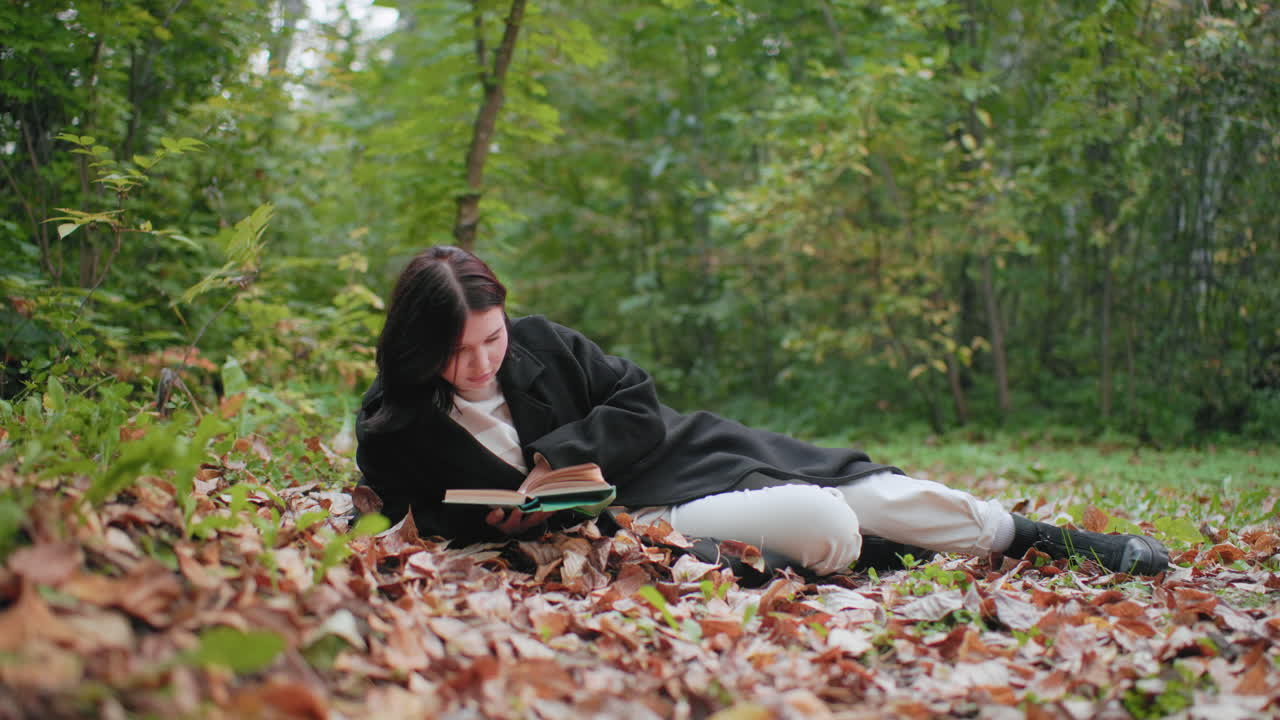 Solitary girl in black coat lies on forest leaf carpet, holding open book and flipping pages with focused attention, enjoying quiet autumn study time, calm outdoor moment surrounded by green foliage