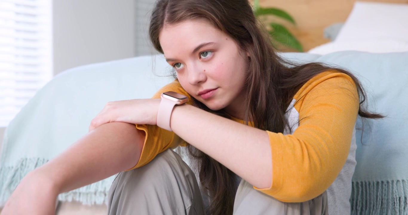 Woman wearing yellow top and pink watch, leaning forward on bed, lifting head toward attentive pose