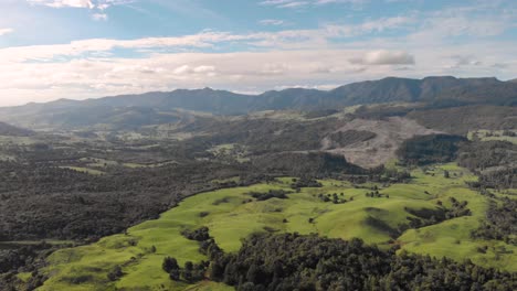 Aerial-view-circling-above-a-green-landscape,-with-hills-and-mountain-ranges-under-a-blue-cloudy-sky