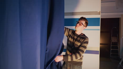 male technician adjusts blue curtain on television studio set during behind scenes broadcast prep, striped backdrop and ladder visible, floor spotlight illuminates cables and equipment