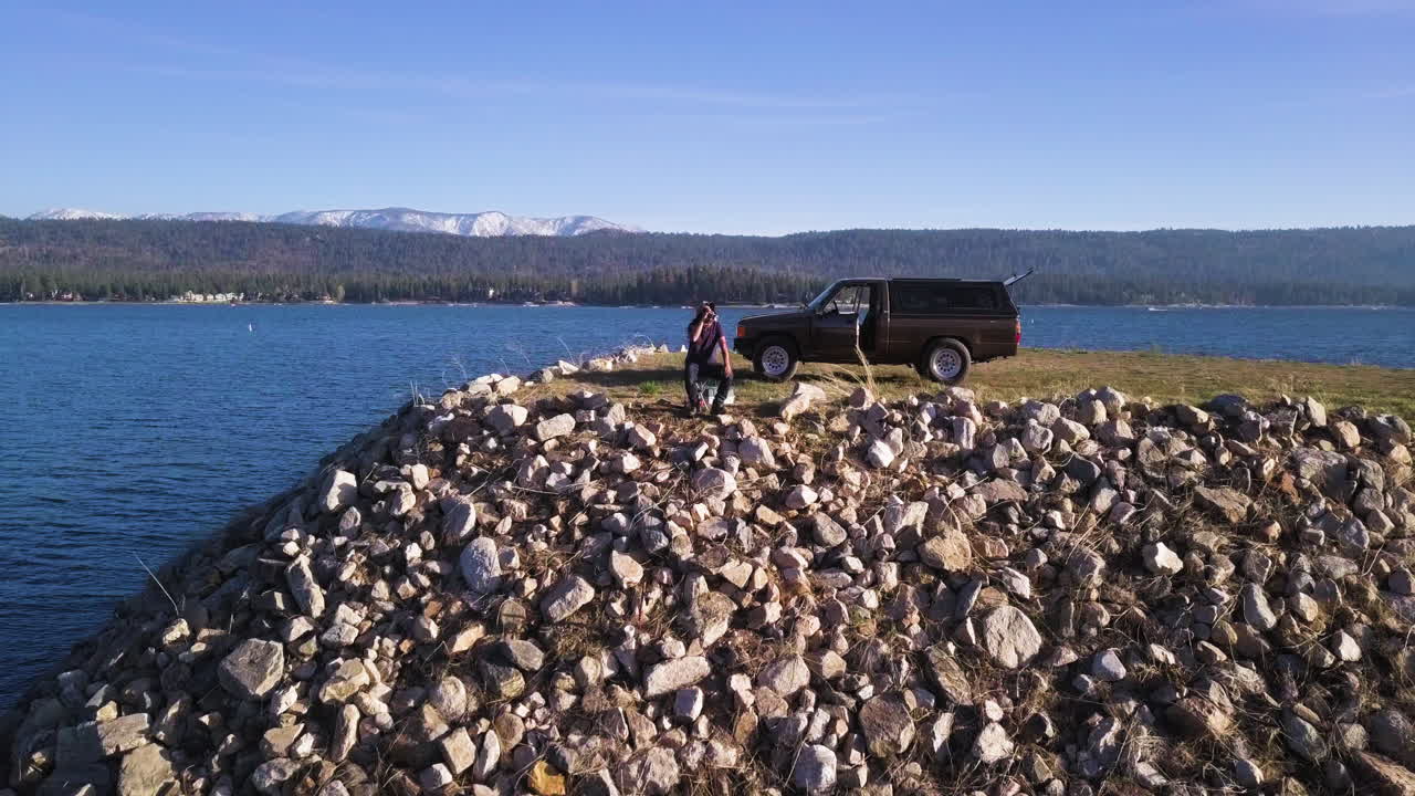 Fisherman enjoying a beer during fishing trip on rocky man made riverbank with truck parked behind him, aerial flying forward