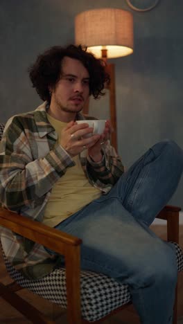 Vertical-video-of-a-brunette-guy-with-curly-hair-brings-a-cup-of-tea-to-his-face-with-both-hands-and-enjoys-a-hot-drink-at-home-on-a-chair-near-the-lamp