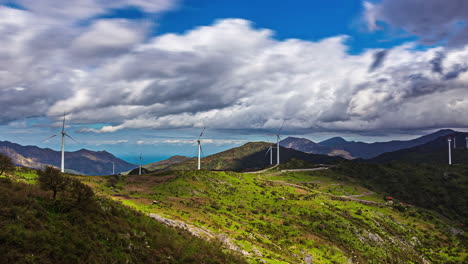 view of rows of white wind mills over a hilly terrain with green vegetation with white clouds passing by on a cloudy day