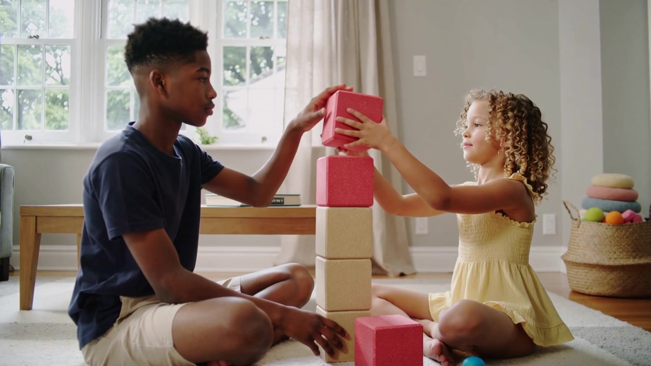 Handing red cube, navy tee boy and yellow-dress girl building beige block tower at home, balancing